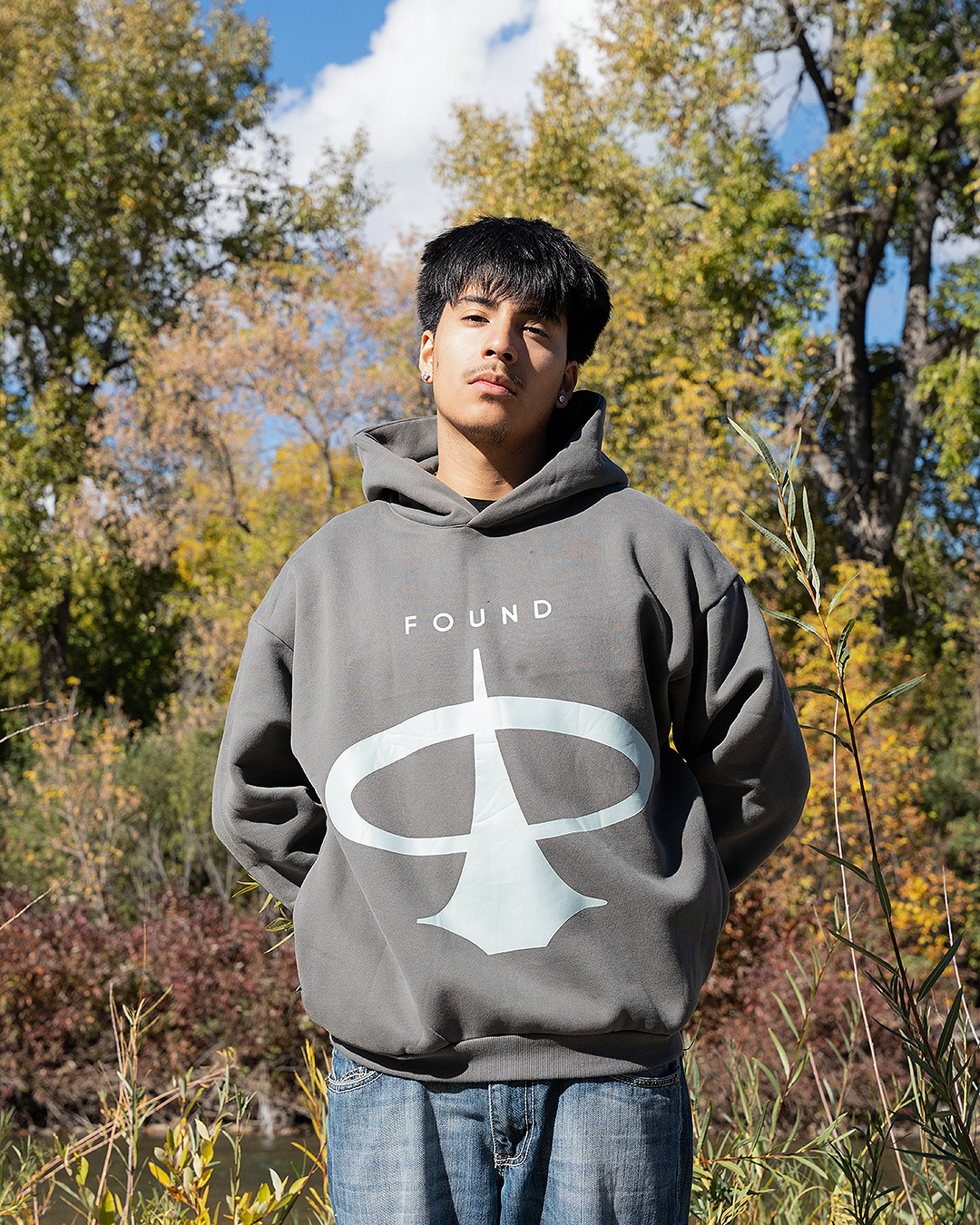 Model facing forward in the FOUND Restoration Hoodie in Ash, full pale-aqua emblem on the chest, autumn trees behind