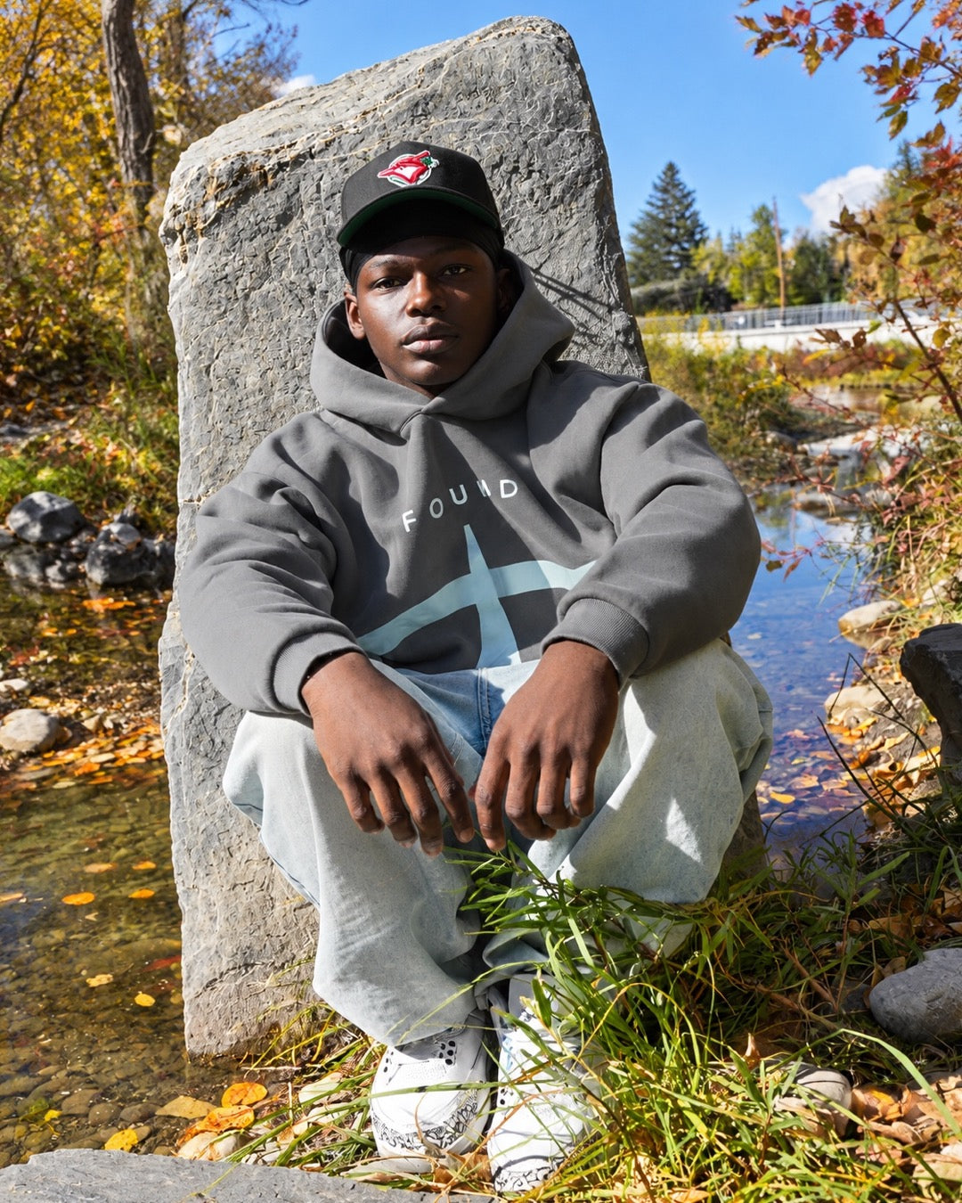 Model seated by a rock in the FOUND Restoration Hoodie in Ash, FOUND wordmark and pale-aqua emblem on the chest