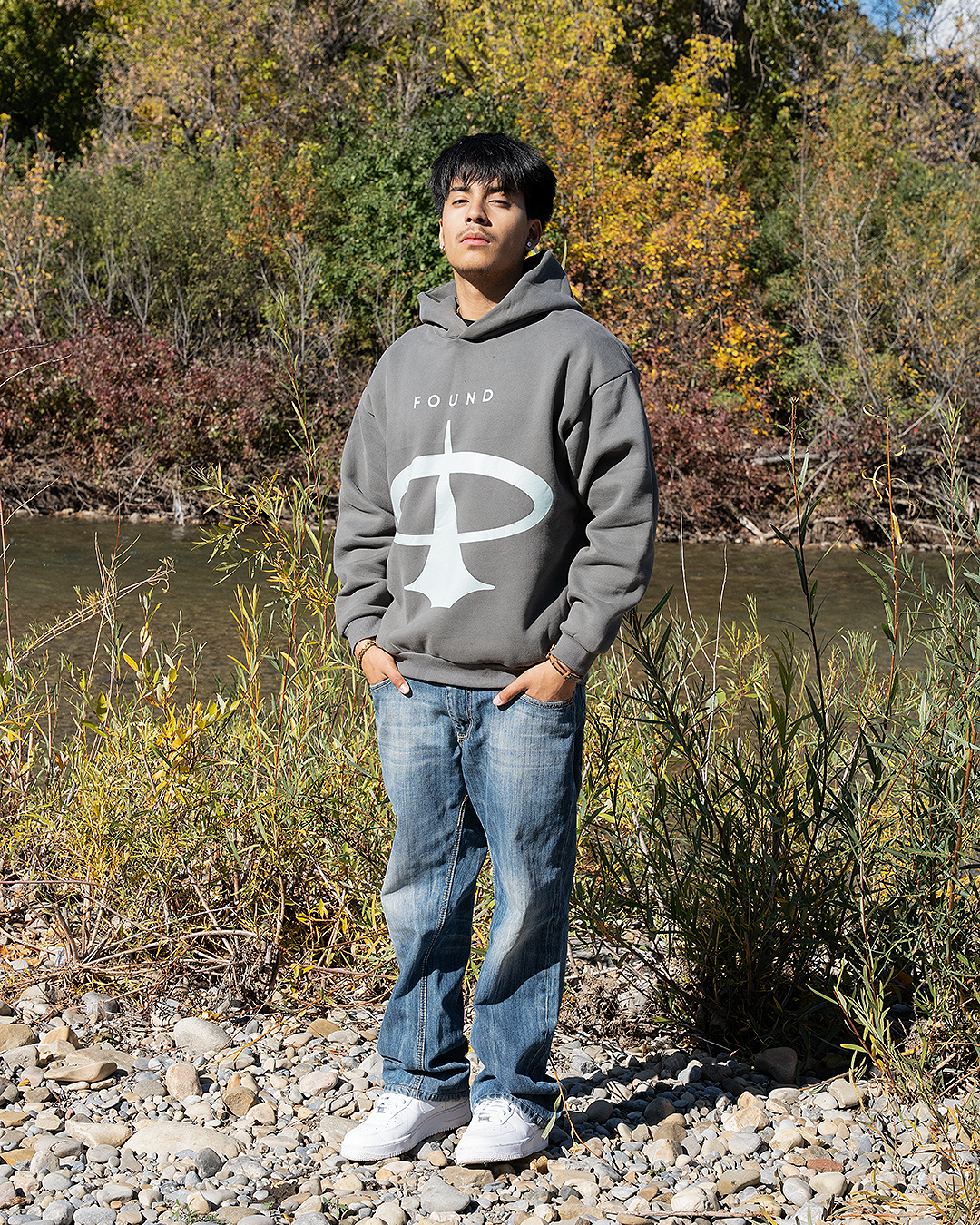 Full-length view of a model in the oversized FOUND Restoration Hoodie in Ash with jeans, by the river
